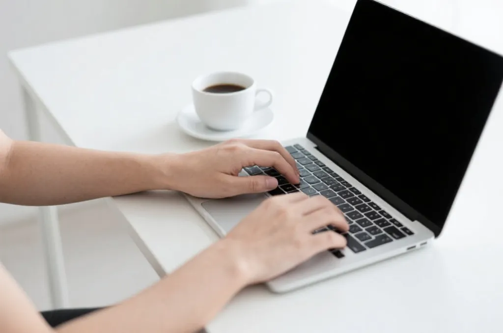 A person typing on a laptop with a cup of coffee next to it, bright minimalist workspace